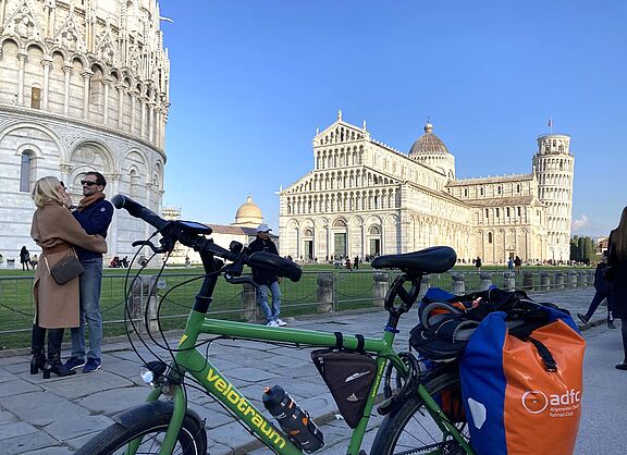 Im Vordergrund steht ein grünes Faltrad mit Gepäcktaschen, darunter eine orange blaue Tasche mit ADFC Logo. Im Hintergrund sind der Domplatz in Pisa mit dem Dom, dem schiefen Turm und ein Teil des Baptisteriums zu sehen, außerdem mehrere Besucher:innen auf der Wiese und auf dem Weg bei blauem Himmel.