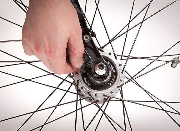 Nahaufnahme eines Fahrradlaufrads mit silbernen Speichen vor weißem Hintergrund. Eine Hand greift von oben in die Speichen und justiert mit einem schwarzen Werkzeug die Nabe in der Mitte des Rads. Rechts ist das Ventil des Reifens zu sehen.